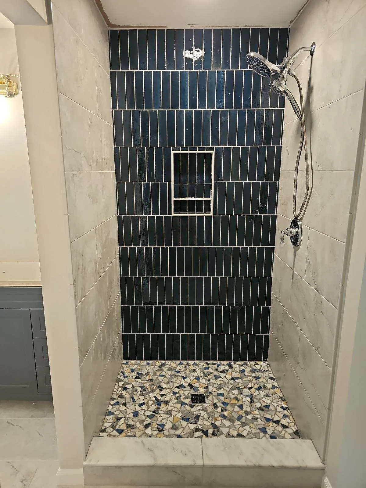 Walk-in shower tiled in deep blue vertical stack-bond tile, framed in marble-look porcelain, over a terrazzo mosaic pan