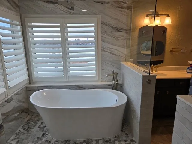 Freestanding soaking tub under plantation shutters with marble-look walls and a hexagon mosaic floor