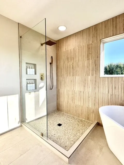 Modern walk-in shower with a glass panel, pebble floor, reeded wood-look tile and brass fixtures