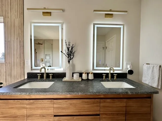 Double vanity with walnut cabinets, a granite counter, backlit mirrors and brass faucets